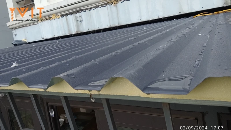 Các đầu vít bắt tại đỉnh sóng tấm tôn xốp màu ghi, mép mái lộ lớp xốp cách nhiệt sau cơn mưa