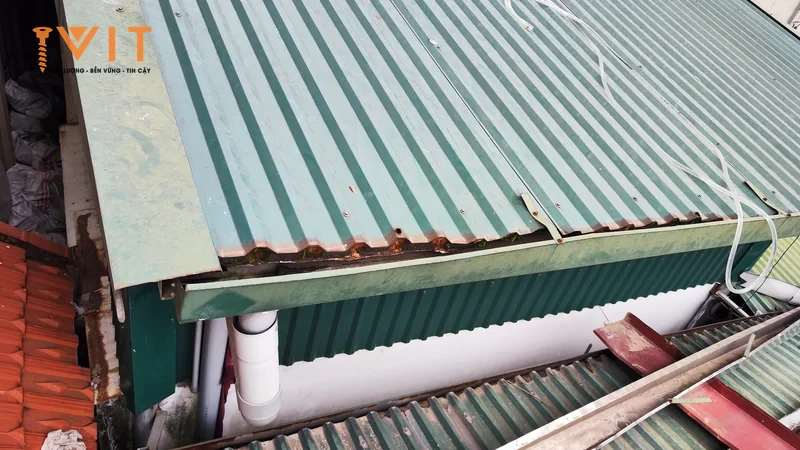 Mép mái tôn sóng xanh bị rỉ sét và vênh lên tại máng xối, cạnh chỗ nối lệch giữa hai tấm tôn của nhà dân cũ