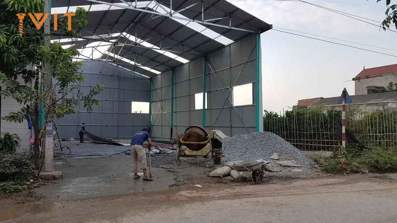 Nhà xưởng khung thép lợp tôn đang hoàn thiện, công nhân đổ bê tông nền cạnh máy trộn và đống đá
