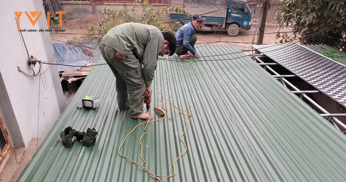 Hai thợ bắn vít lợp mái tôn sóng màu xanh bằng máy bắn vít, dây điện trải dọc theo tấm tôn