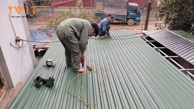 Thợ bắn vít tự khoan trên mái tôn sóng màu xanh, dây máy bắn vít trải dọc mặt tôn