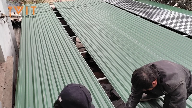 Hai thợ thi công đang lợp mái tôn sóng màu xanh, một người cúi sát mép tấm tôn trên mái nhà