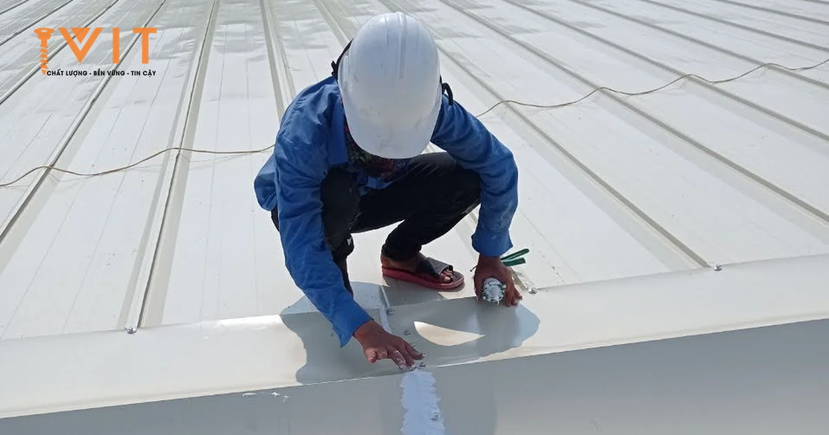 Vít bắn úp nóc bắt thành hàng dọc đỉnh mái tôn màu kem, thợ đội mũ bảo hộ trét keo kín mối nối hai tấm úp nóc